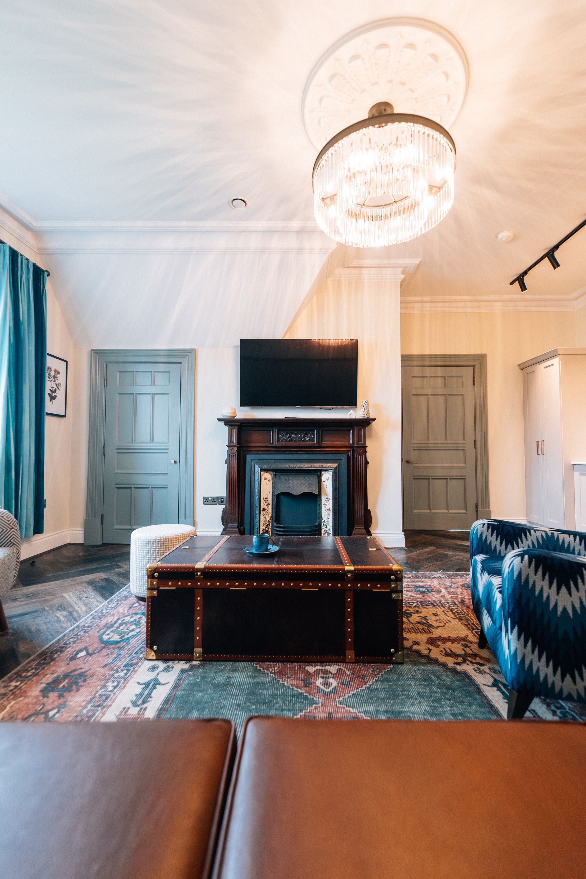 Living room with fireplace, TV, and trunk coffee table. Two blue armchairs and a large chandelier are also visible.