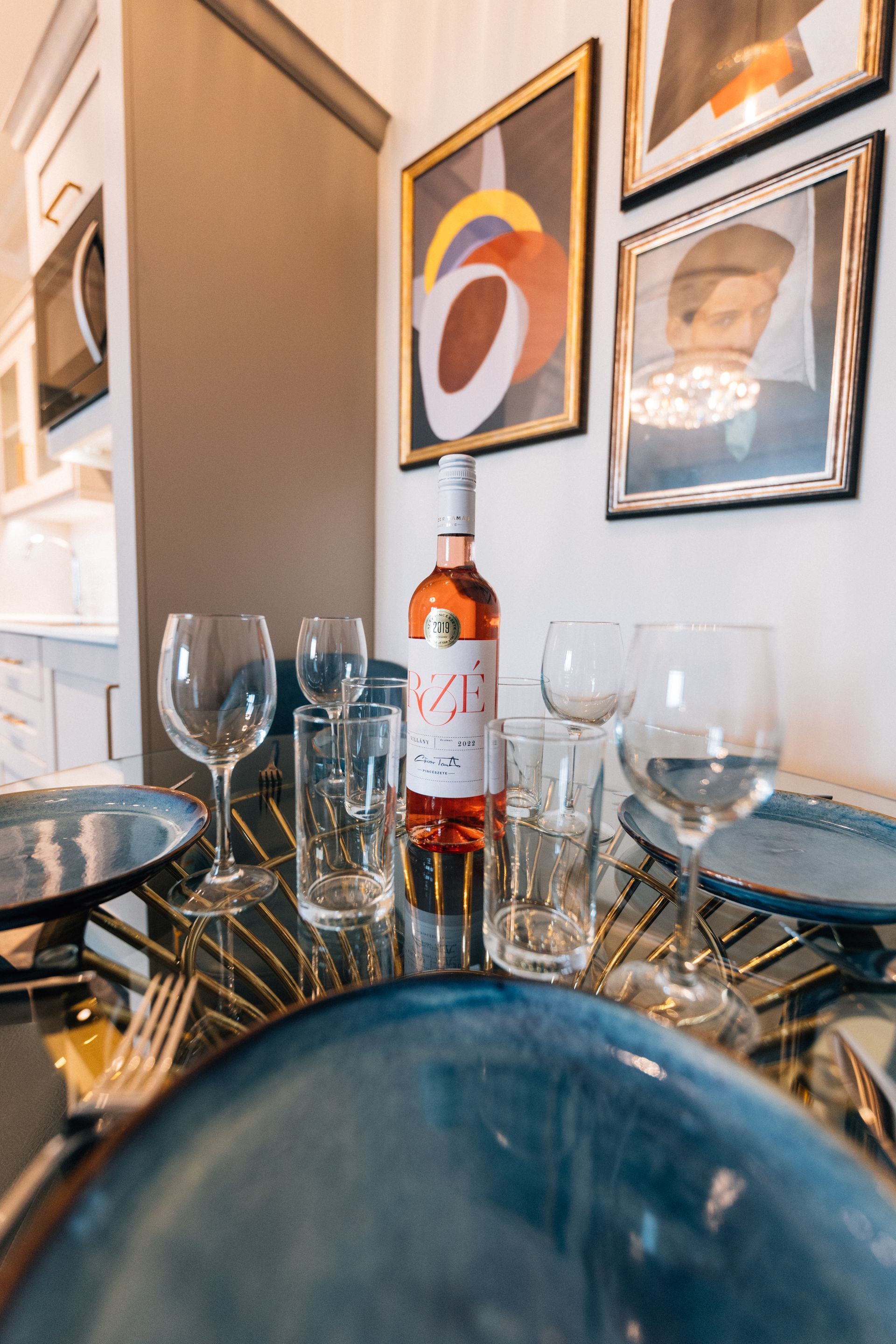 A table set for a meal with blue plates, wine glasses, and a bottle of rosé wine; framed art hangs on the wall behind.