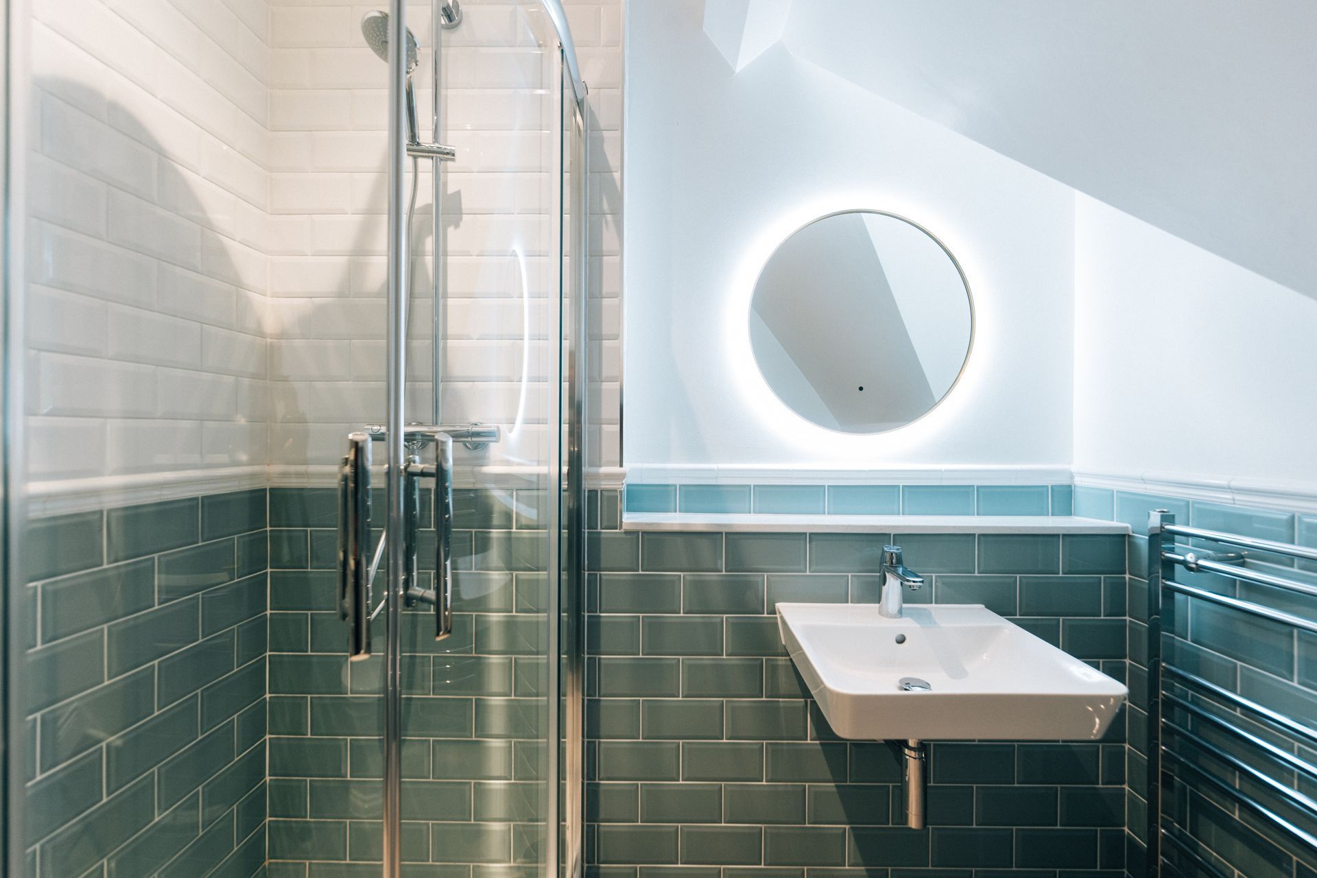 A modern bathroom featuring a glass-door shower, sink, and a backlit circular mirror; the walls are tiled in teal and white.