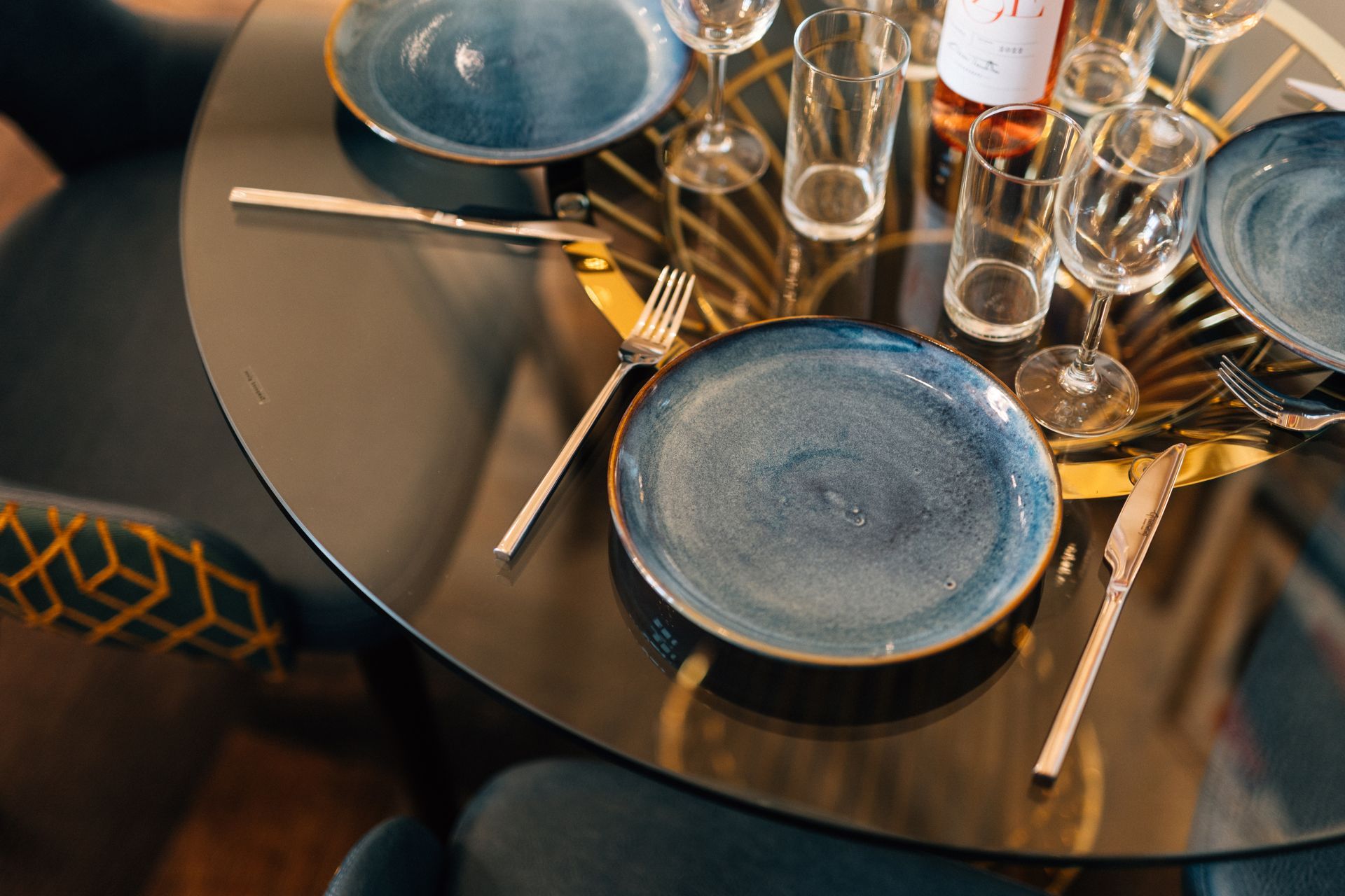 Round table set for dining with blue plates, glasses, silverware, and a bottle of rosé.