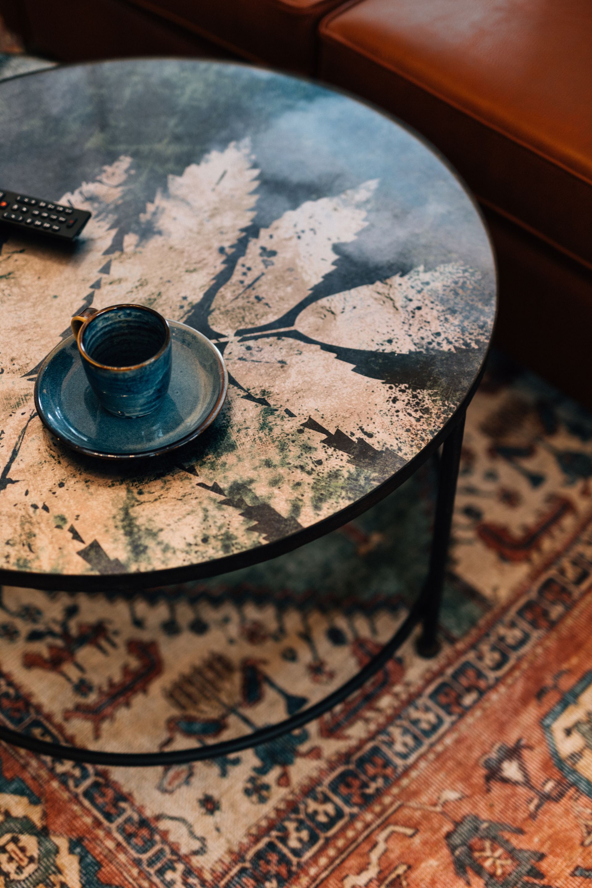 Round coffee table with a leaf pattern, a teal cup and saucer, and a remote.  