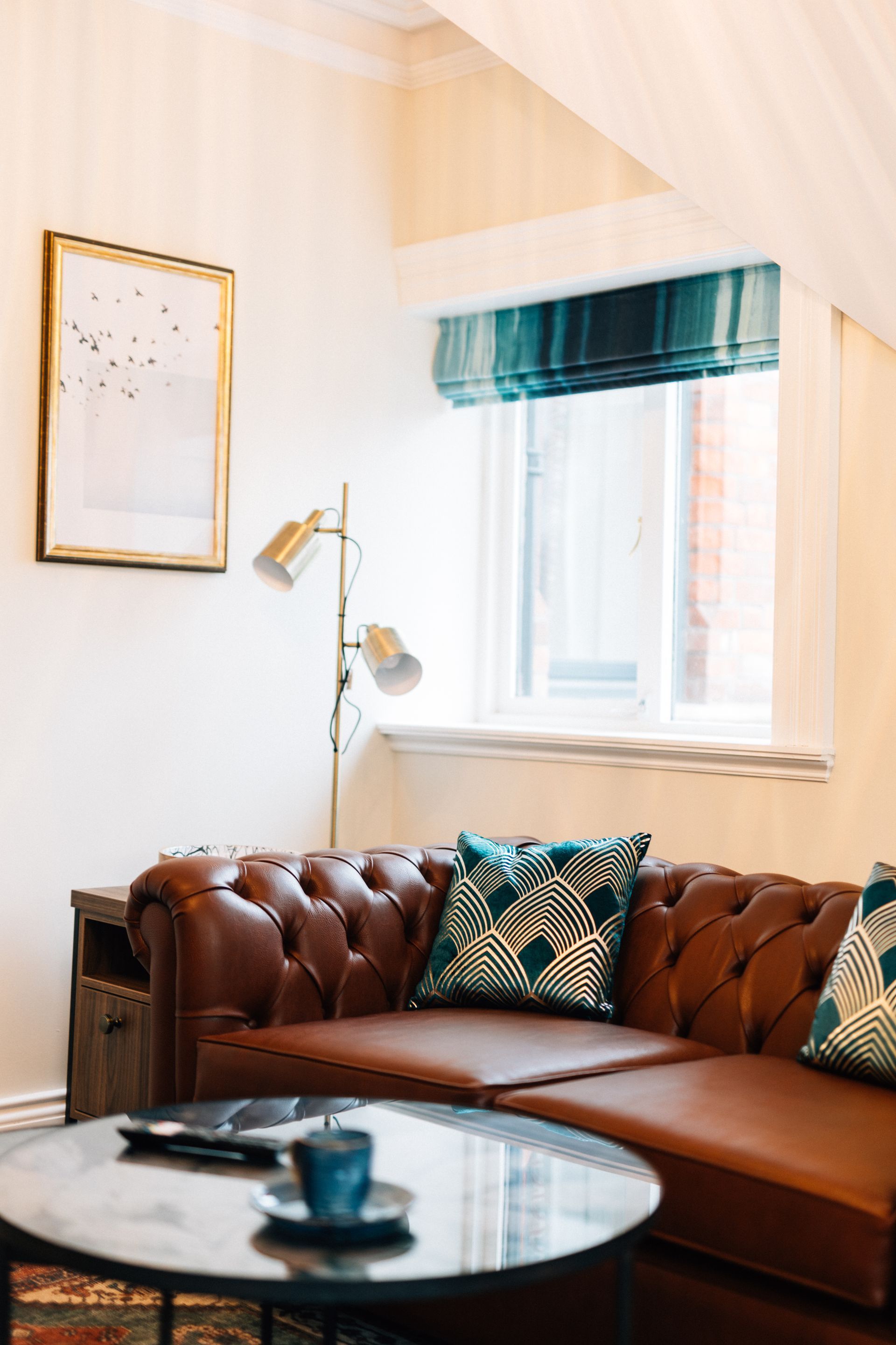 A cozy living space with a brown leather sofa, a round coffee table, and a window with a blue and white shade.