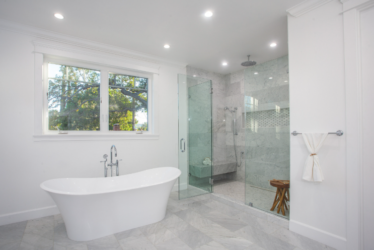 White bathroom with a freestanding tub, glass shower, and large window.