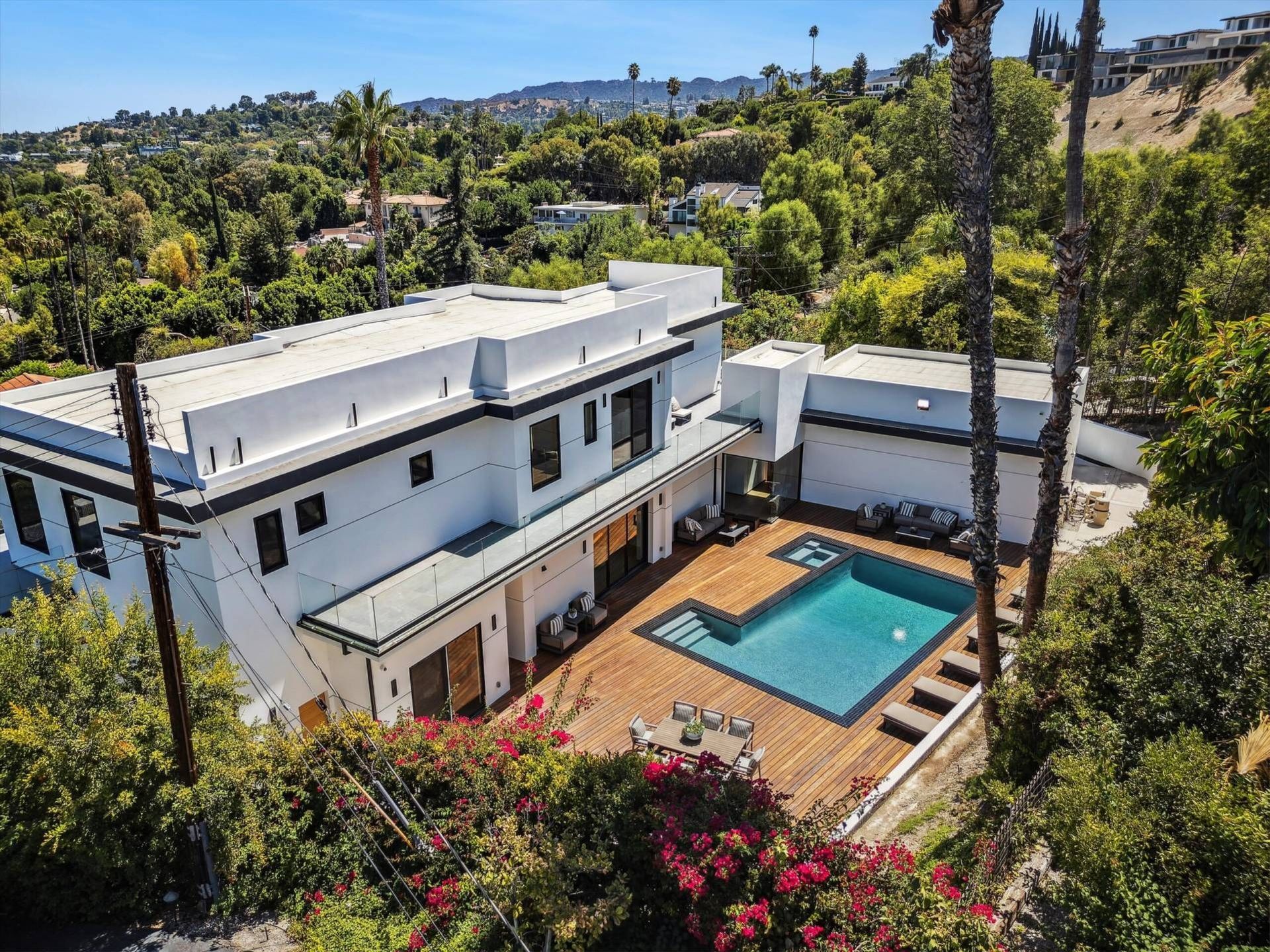 Modern white house with a pool on a wooden deck surrounded by lush green trees on a hillside.