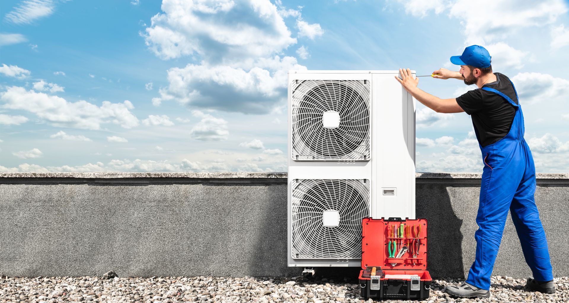 HVAC technician in blue overalls and cap, installing an AC unit on a rooftop.