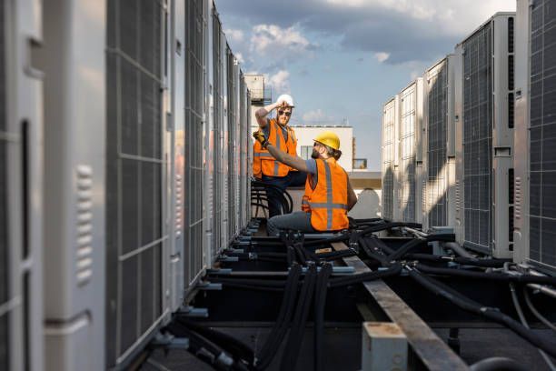 Two HVAC technicians on a rooftop. They are inspecting machinery while wearing hard hats and orange vests.