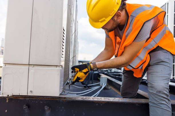 Construction worker kneels, fixing wires on a rooftop, wearing a yellow hard hat and orange vest.
