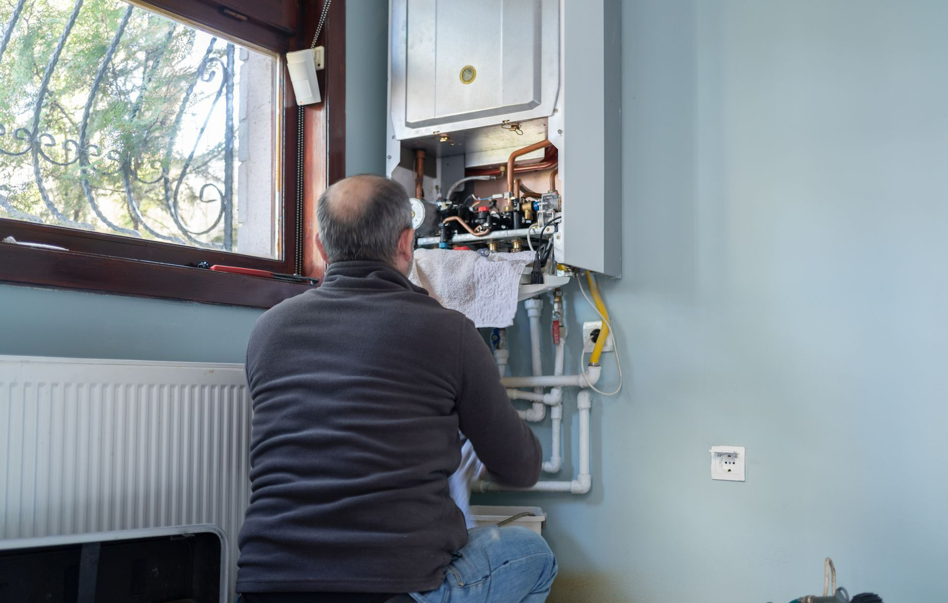 Man in blue jeans kneeling by a wall-mounted furnace, likely repairing it, near a window.