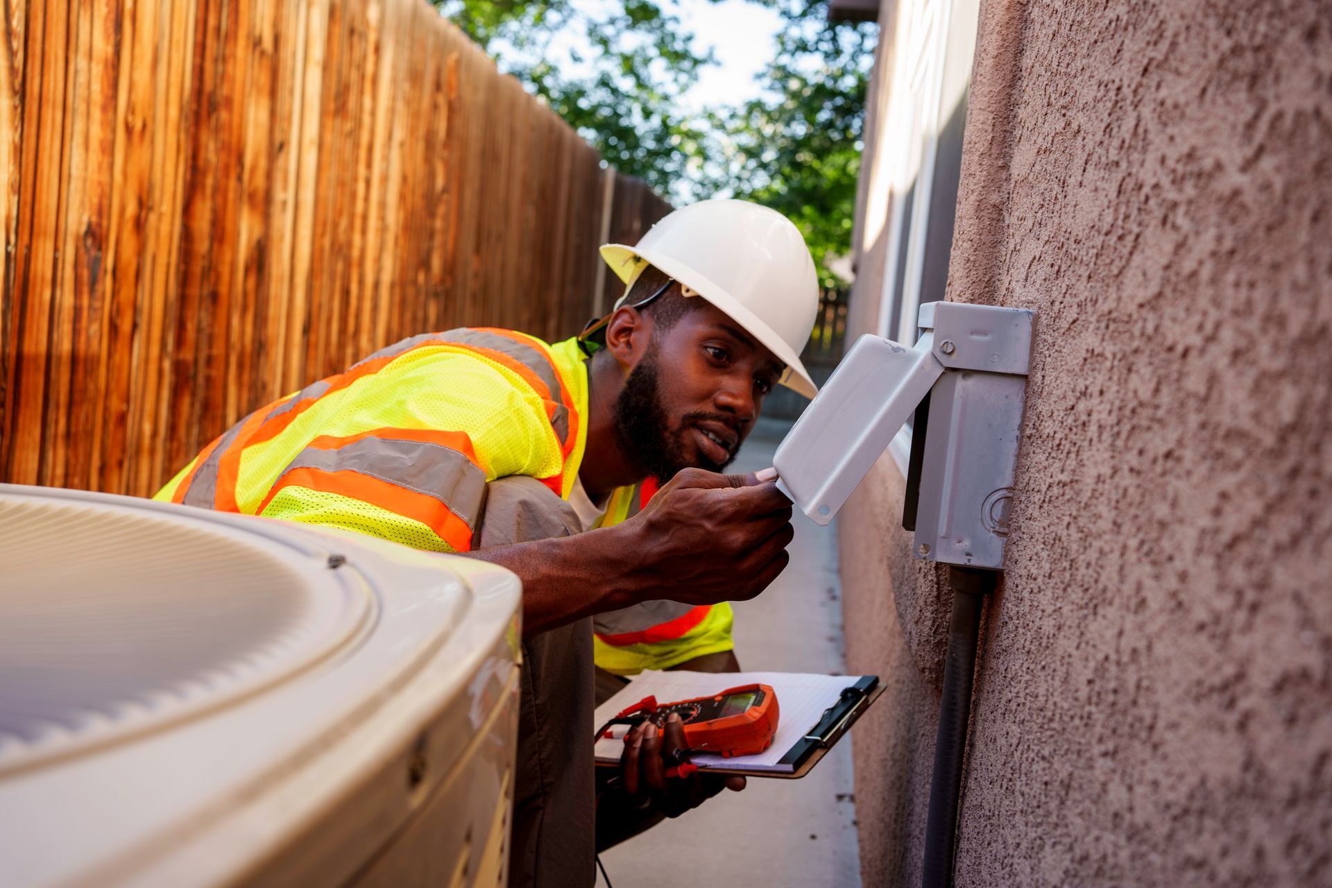 Black HVAC technician inspecting a unit, wearing a hard hat and vest, near a wall and fence.