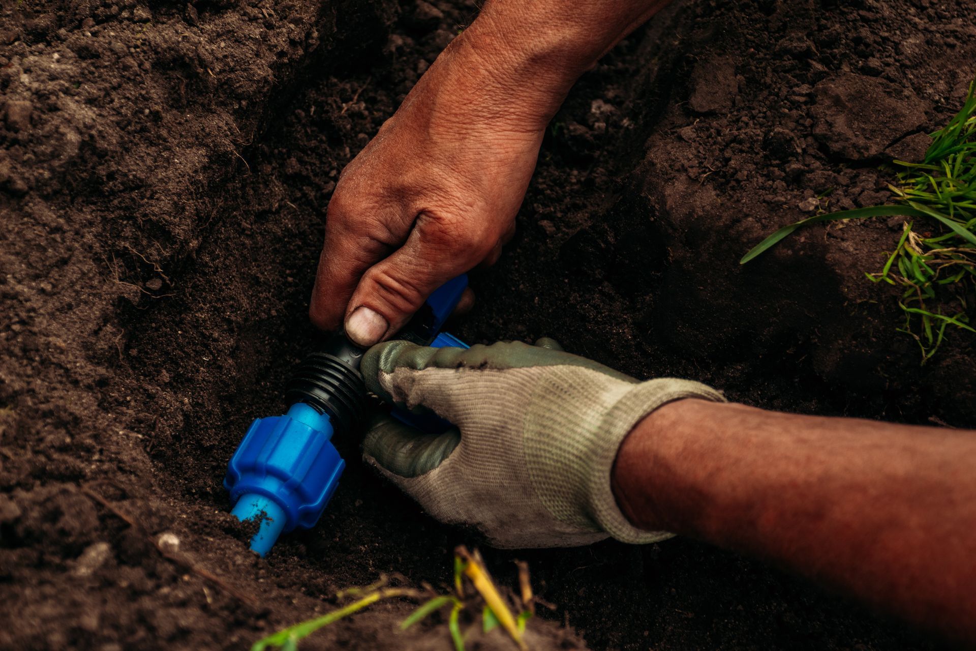 Hands installing a blue irrigation fitting in a dug trench, one wearing a work glove.