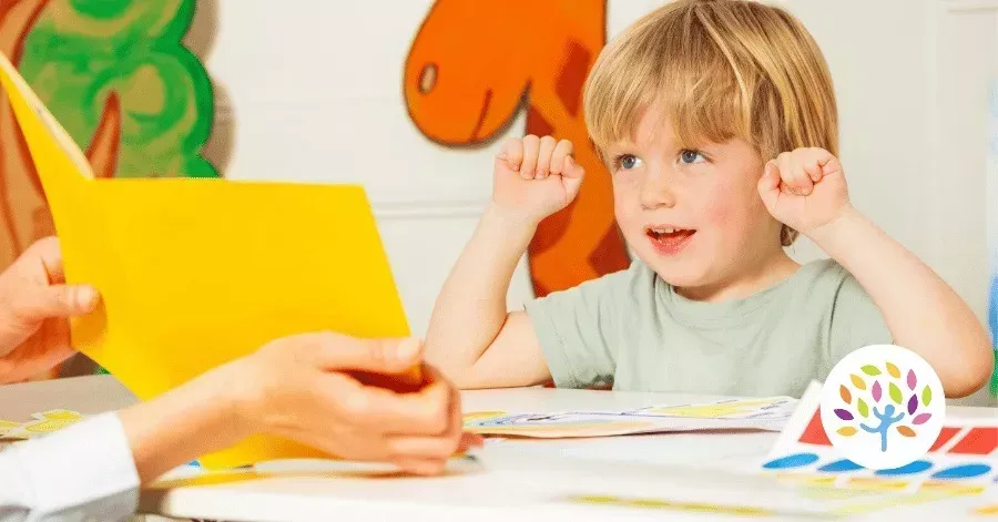 Child raising hands at a table while someone holds a yellow folder in a colorful classroom setting
