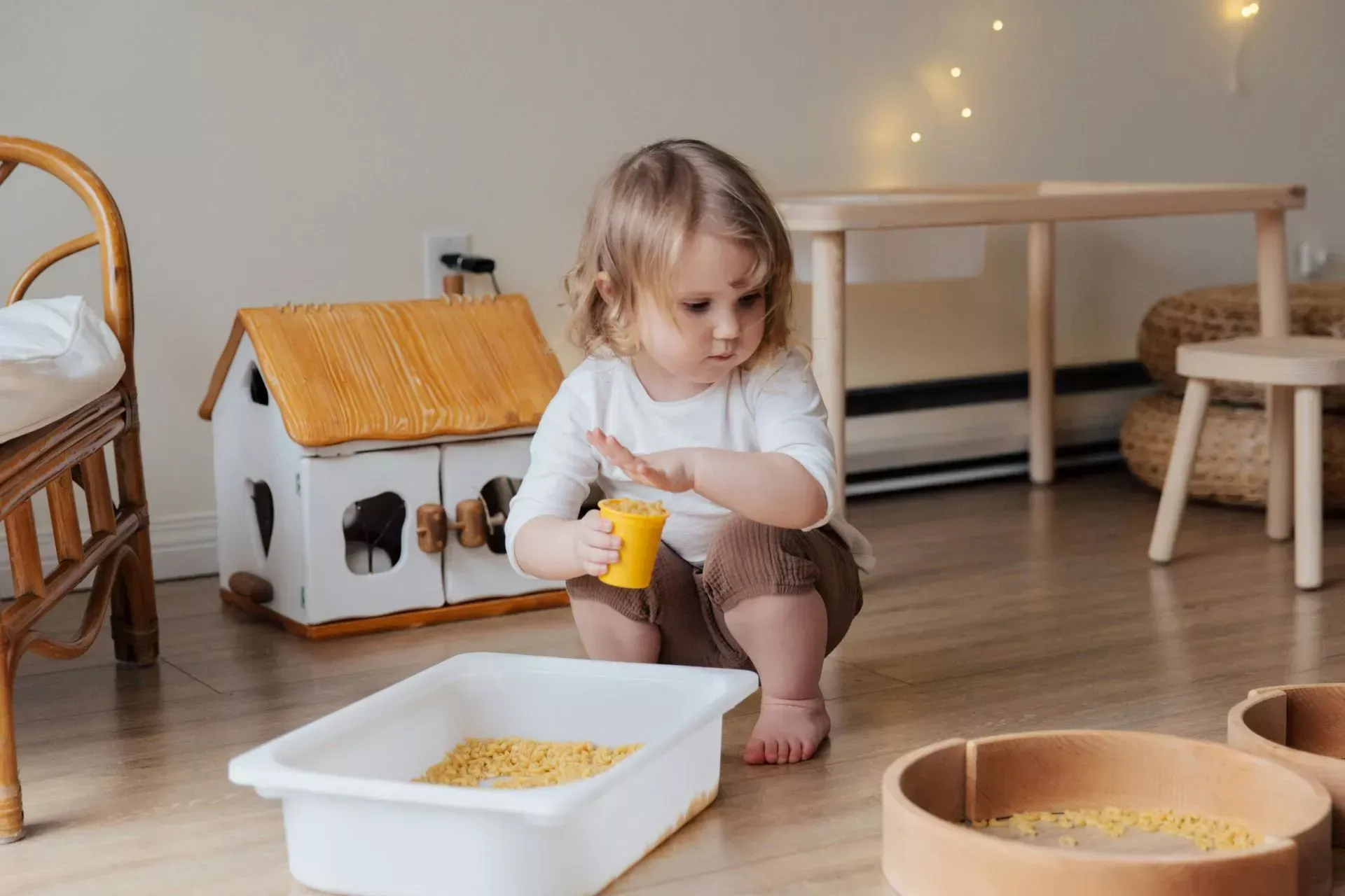 Child playing with yellow toy in a cozy playroom with wooden floors, bins, and soft lights
