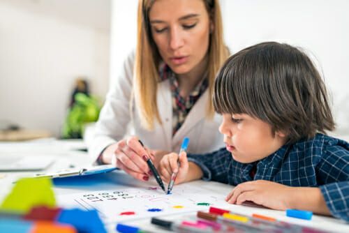 Woman in white coat helping a child color a worksheet with markers.