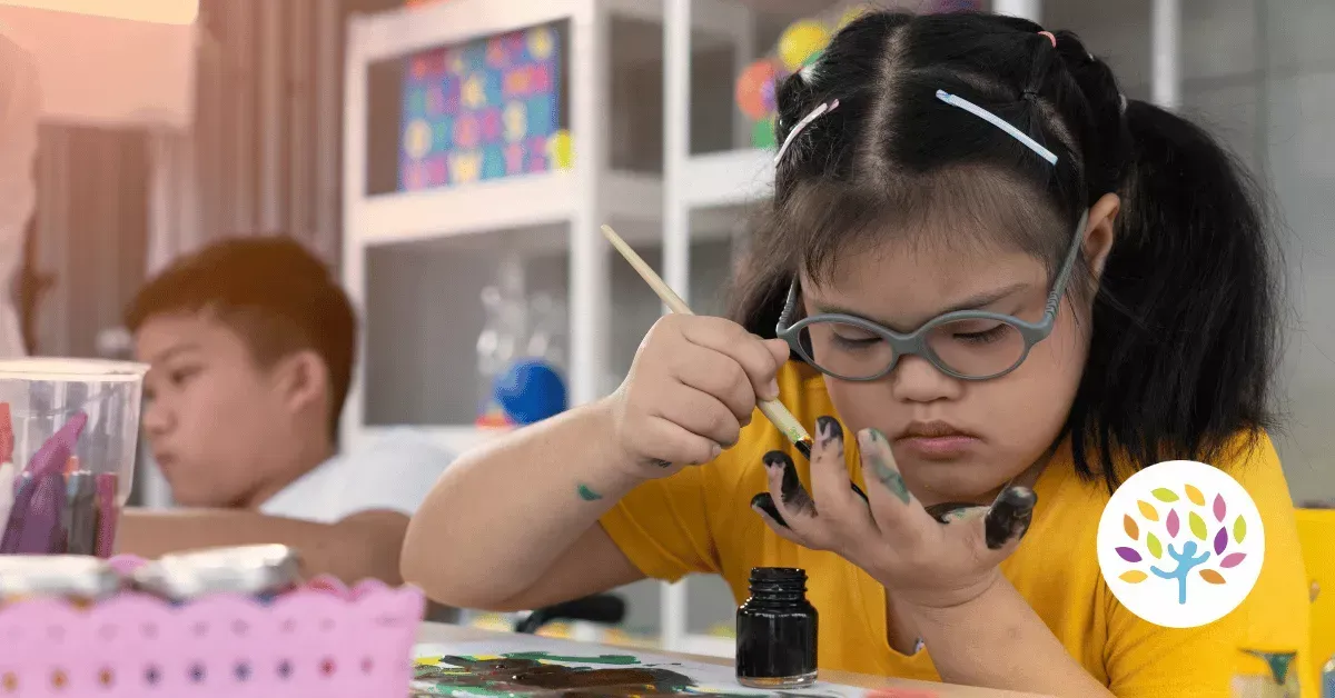 A student with glasses in a yellow shirt carefully paints a small object with a brush at a table.