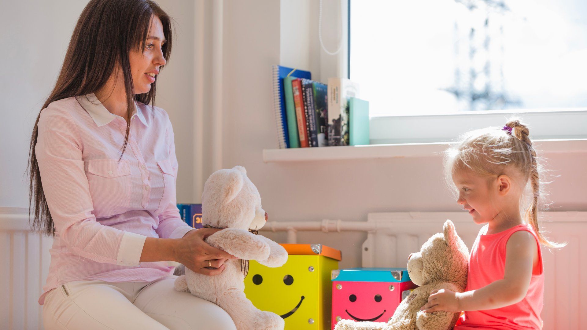 Woman and child playing with teddy bears near a window.