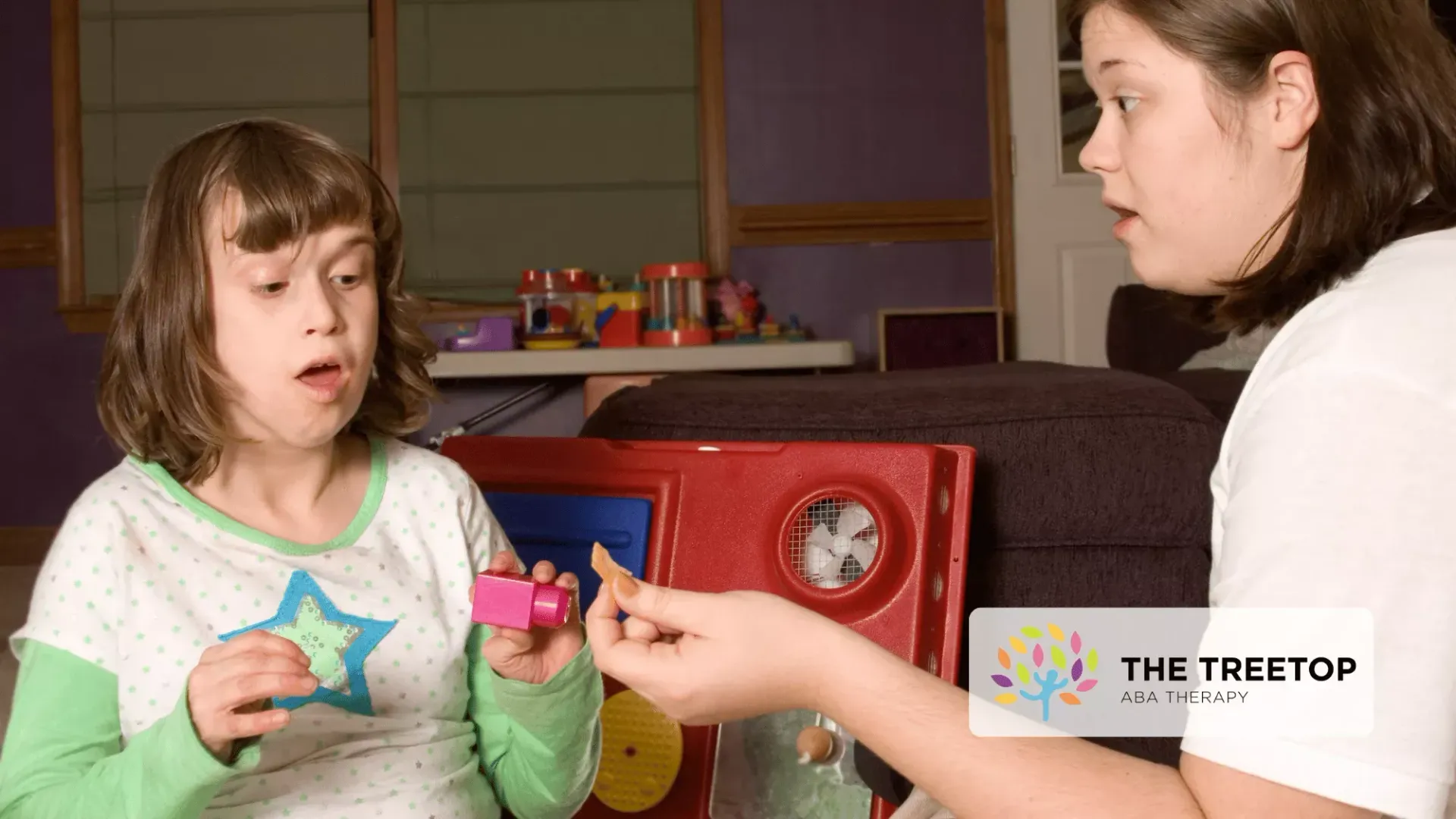 A therapist interacts with a child holding a pink block in an indoor therapy setting.
