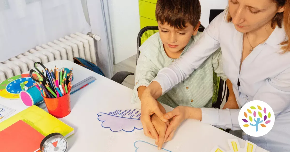 An adult guides a child’s hand while drawing clouds and rain on a piece of paper at a desk filled with school supplies.