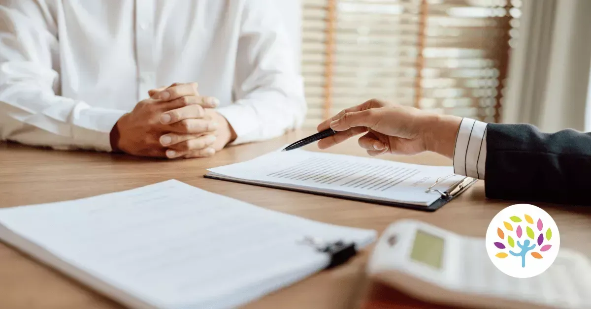 Two people seated at a wooden desk with documents and a calculator, one person pointing to a paper during a consultation.