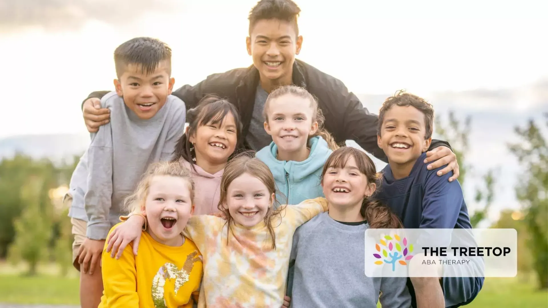 A group of eight smiling children pose outdoors in a sunny field, some with arms around each other.