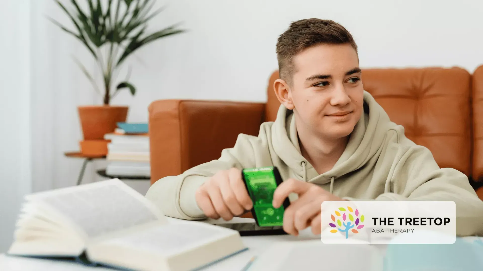 A student in a beige hoodie sits at a table with an open book and a small green device, looking off to the side.