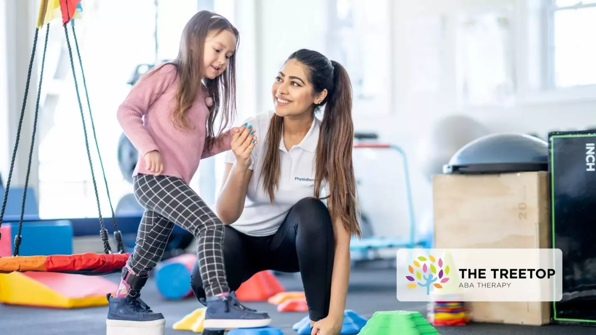 A therapist kneeling in a gym encourages a child as they step onto a platform during an exercise session.