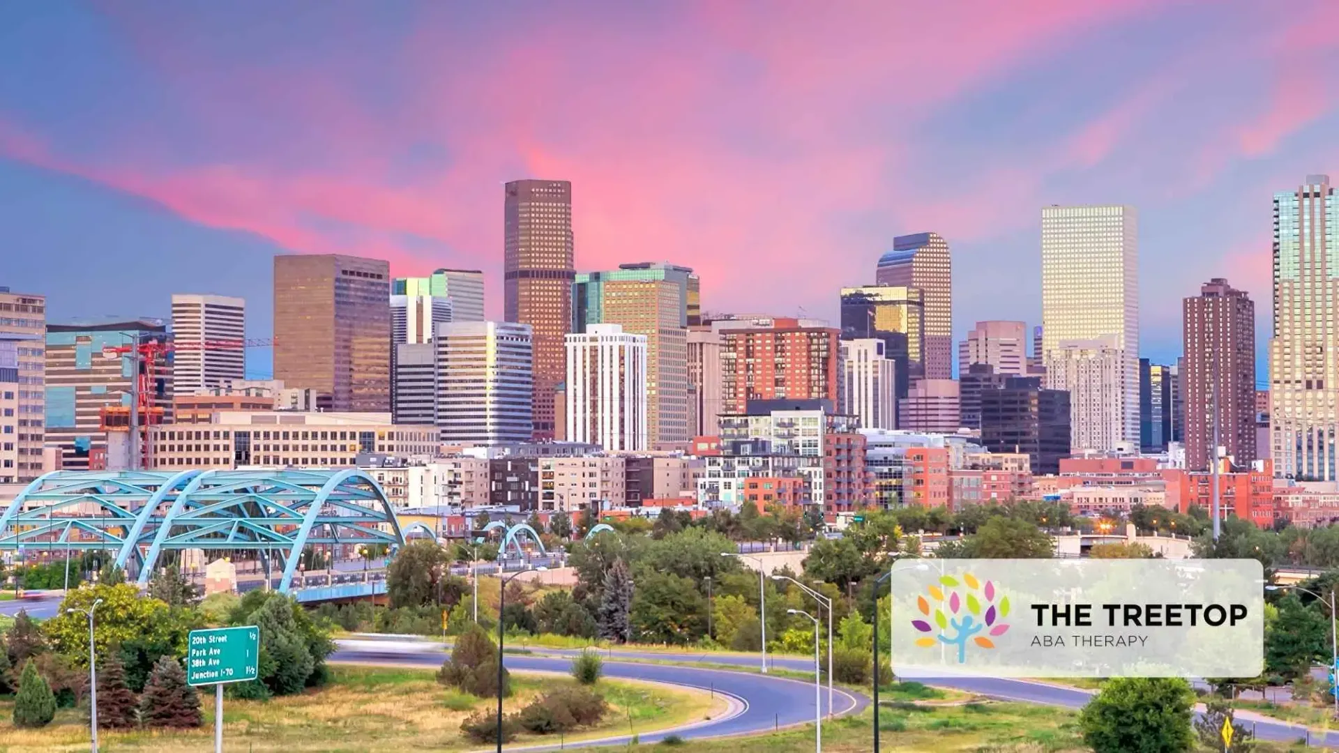 City skyline at sunset with a pink sky, lit buildings, and a park sign reading “The Treetop” in the foreground