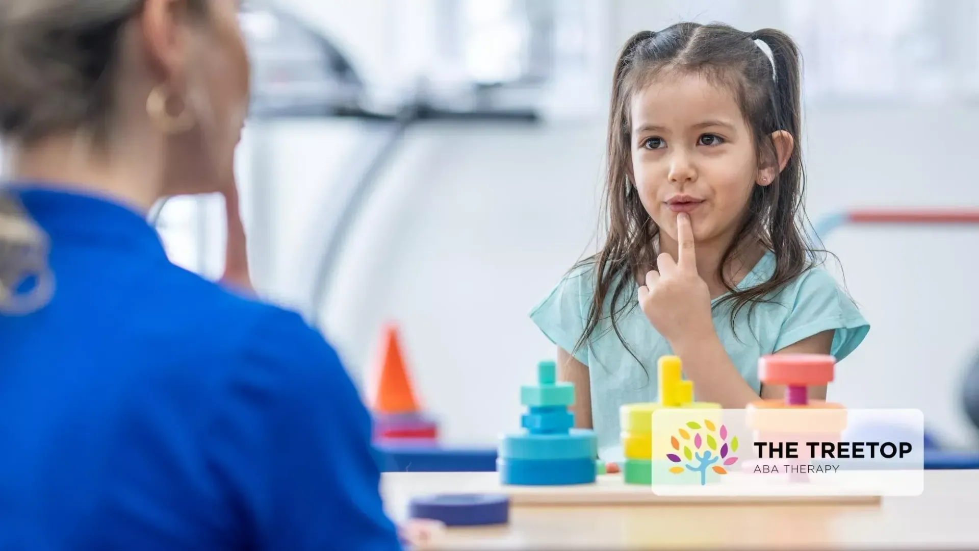 Child thinking at a desk with a teacher nearby, using colorful learning blocks in a classroom