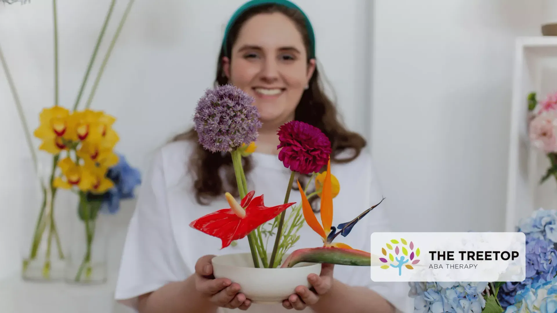 A smiling person holds a decorative arrangement of colorful flowers in a white bowl, set in a bright, floral studio.