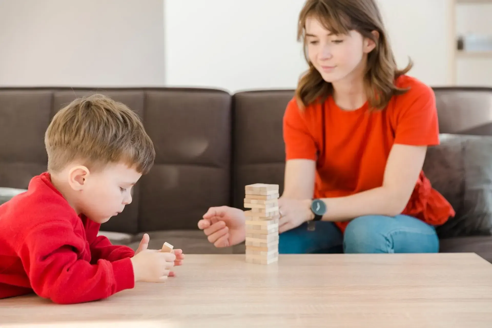 A person in a red shirt sits on a brown sofa, playing a wooden block stacking game with a child on a wooden table.