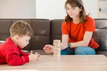 A person in a red shirt sits on a brown sofa, playing a wooden block stacking game with a child on a wooden table.