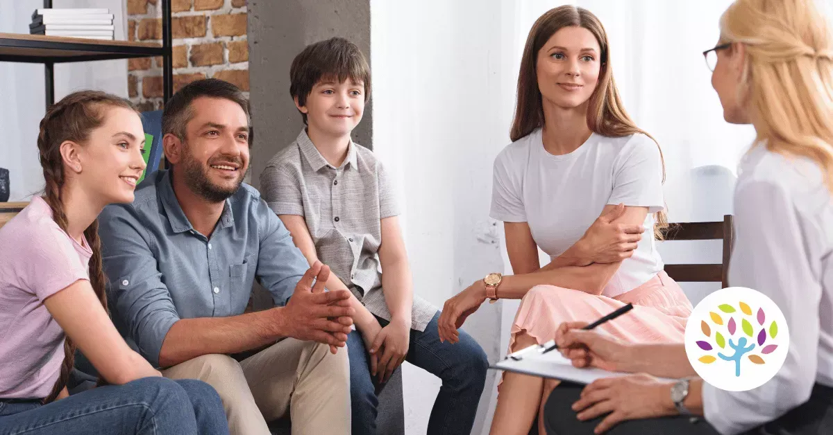 A family of four participates in a counseling session, sitting together in a room and talking to a professional.