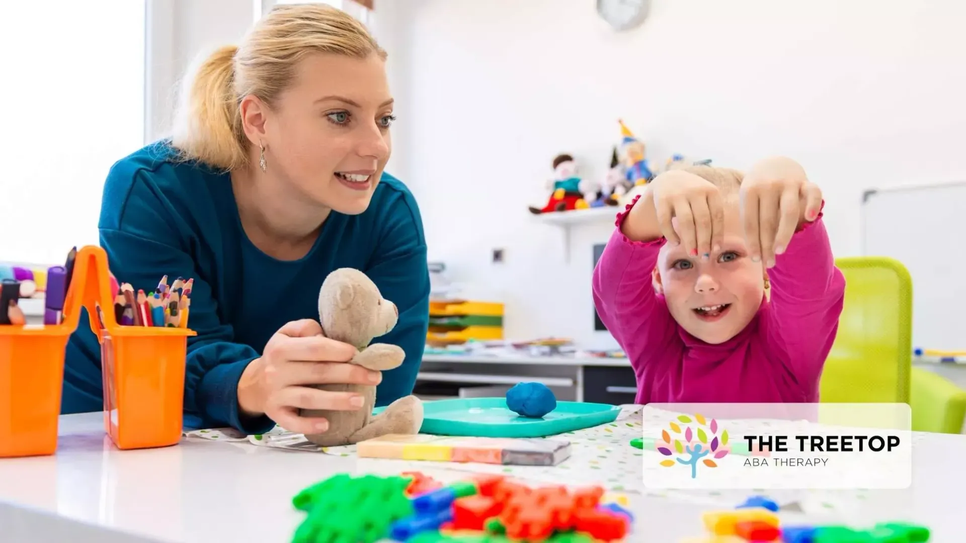 An instructor sits at a table with a child, using a small stuffed bear to engage in play-based learning.