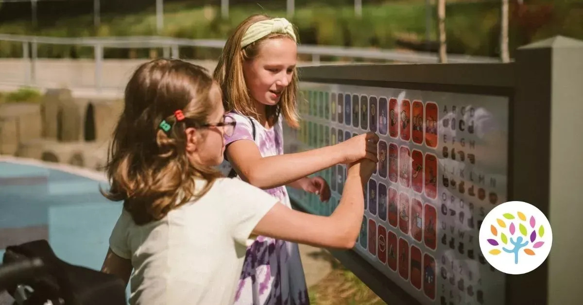 Two children stand outdoors at a park, pointing at and interacting with a grid of colorful symbols on an educational sign.