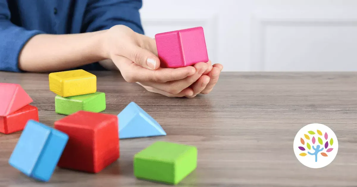 A person holds a pink wooden cube above a table covered with various colorful geometric wooden blocks.