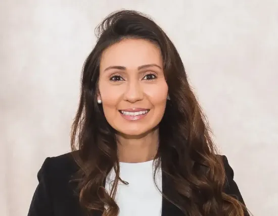 A person with long brown hair, wearing a white shirt and black blazer, smiling at the camera against a neutral background.