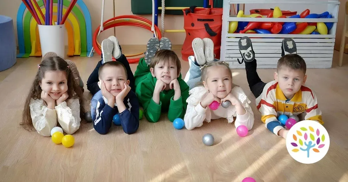 Five children lie on their stomachs on the floor in a colorful playroom, resting their chins on their hands.