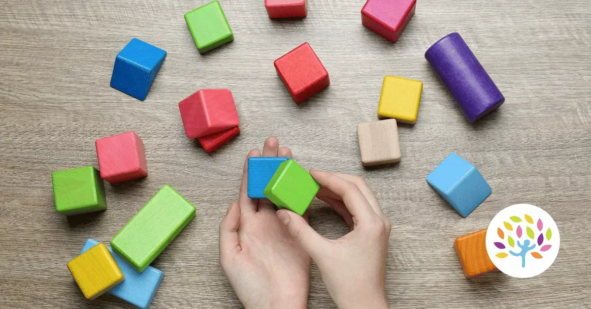 Hands hold a green block among various colorful wooden geometric shapes scattered on a light wooden surface.