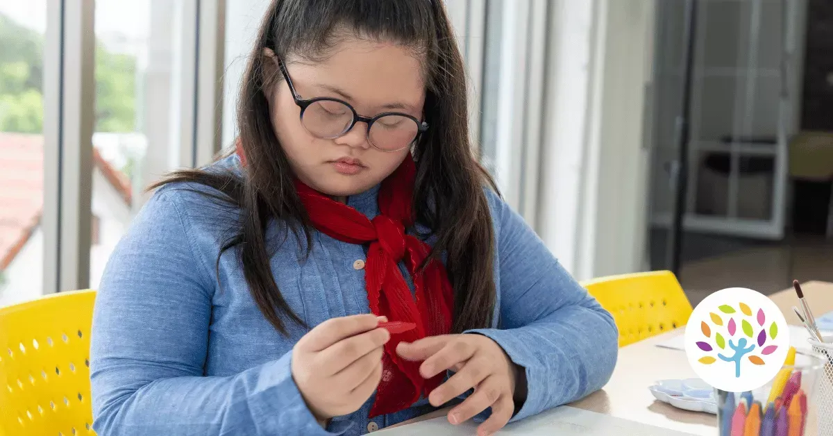 A person with Down syndrome wearing glasses and a red scarf, sitting at a table while focused on an art activity.