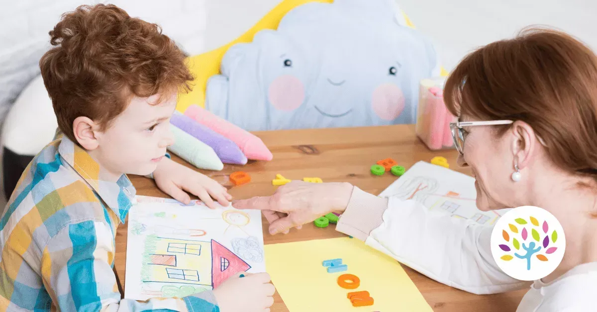 A teacher points to a drawing while working with a young student at a wooden desk with learning materials and a cloud pillow.