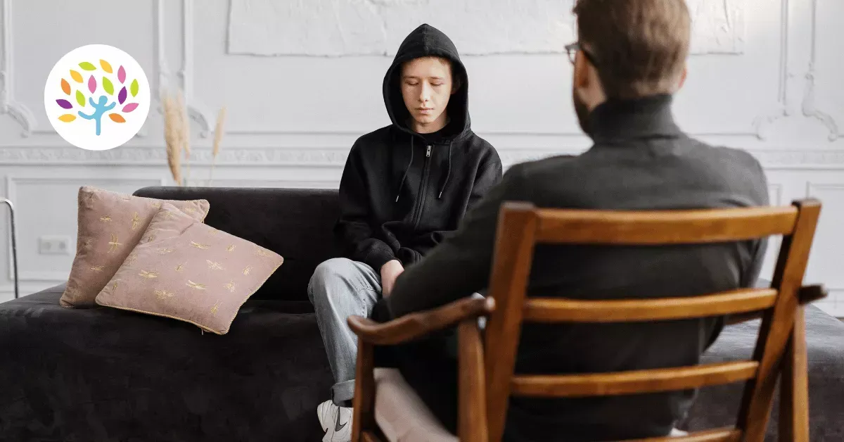 A person wearing a black hoodie sits on a sofa, facing a professional who sits in a wooden chair during a consultation.