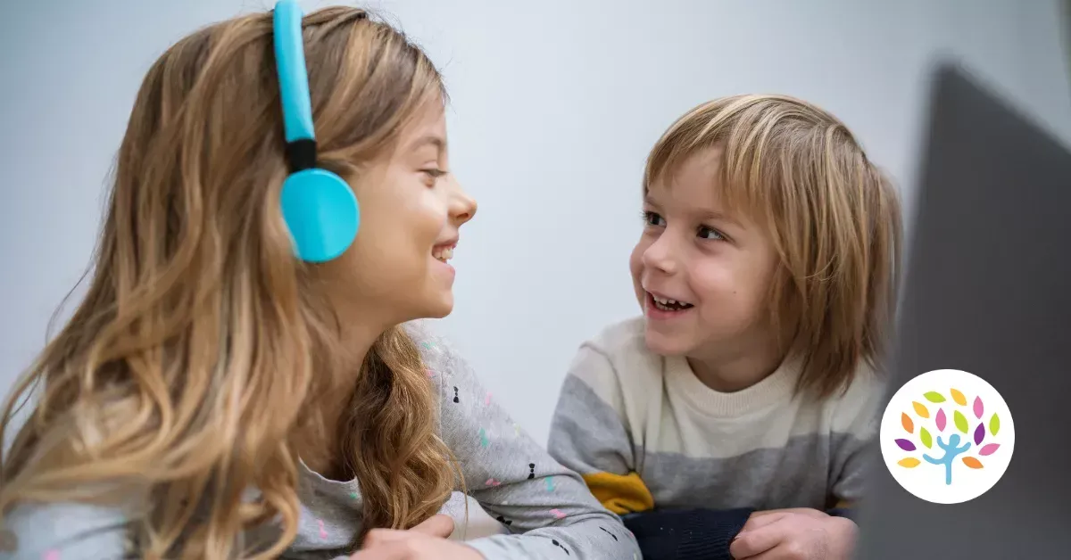 Two children smiling at each other while looking at a laptop, one wearing blue headphones. A colorful tree logo is visible.