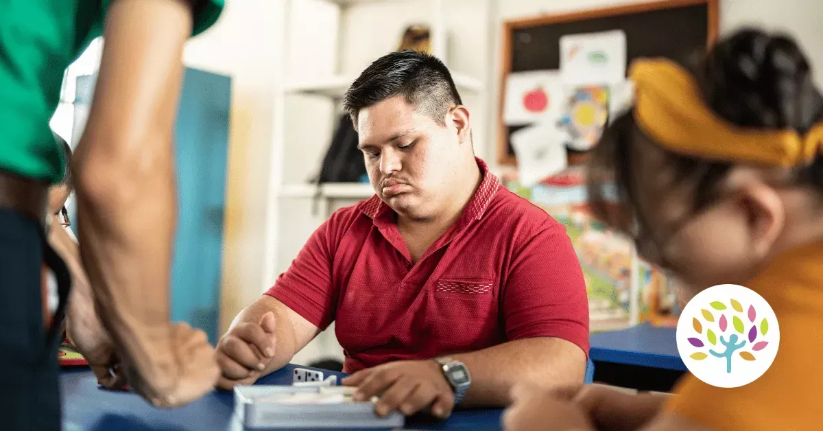 A person in a red polo shirt sits at a desk with others, focused on an object in front of them in a classroom setting.