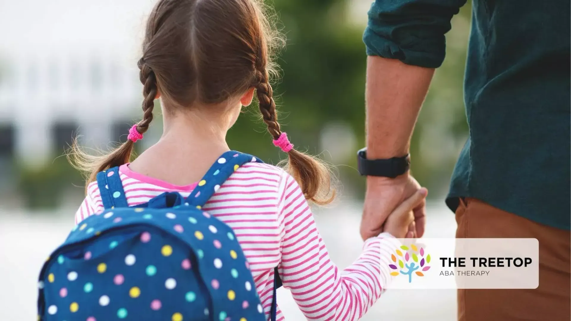 Child with blue polka-dot backpack holding an adult’s hand outdoors.