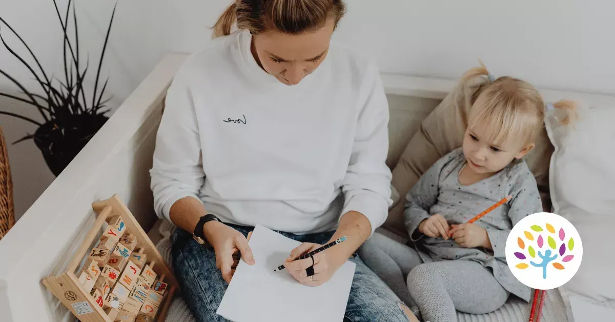 A person writing on paper next to a toddler holding a pencil, seated together on a sofa.