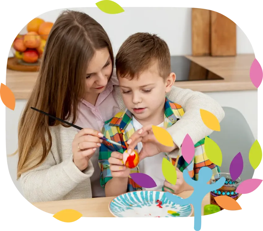 A person helps a child paint a small egg at a wooden table in a kitchen.