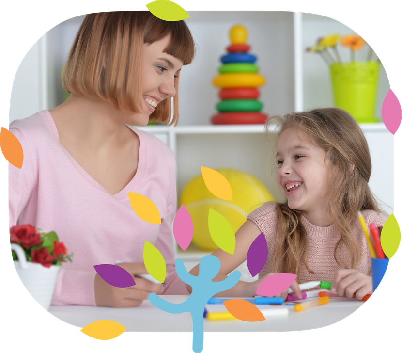 A person and a child smiling while drawing with markers at a table in a brightly lit room with a toy pyramid behind them.
