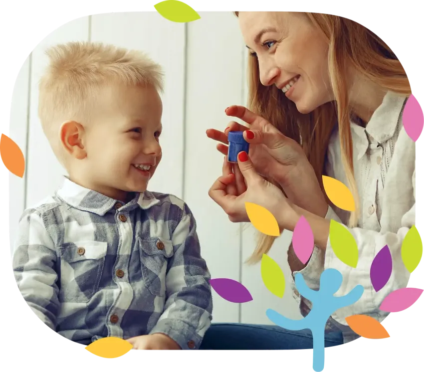 A caregiver smiles while holding a small blue object for a child to look at, set against a background with colorful leaves.