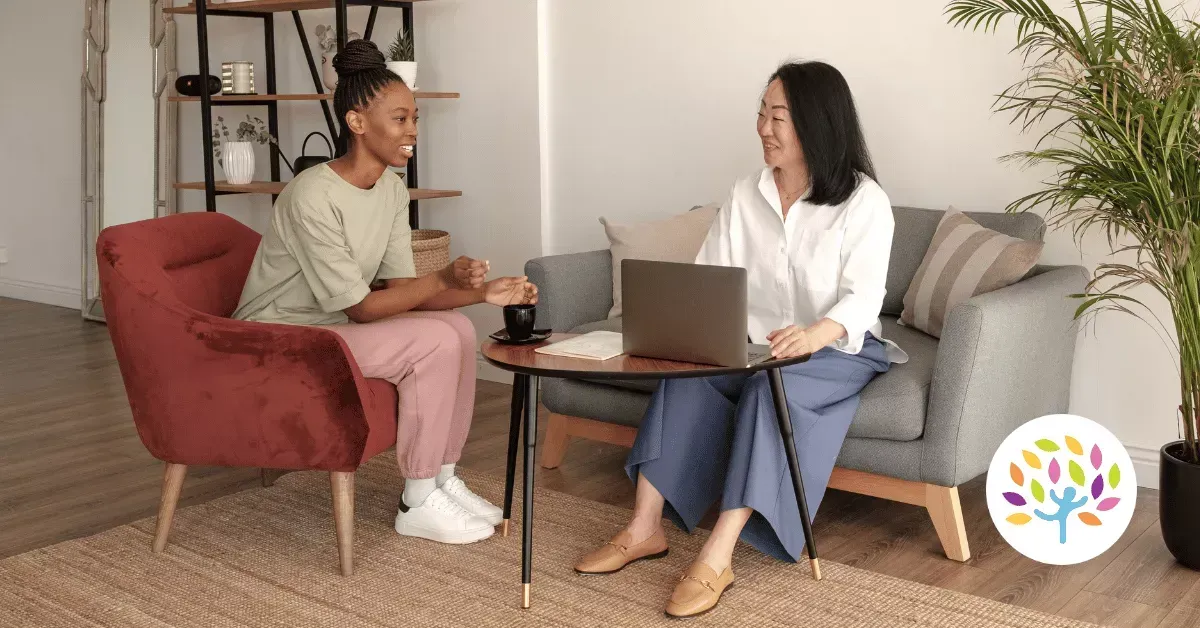 Two people sit in a living room, talking while one uses a laptop on a coffee table between them.