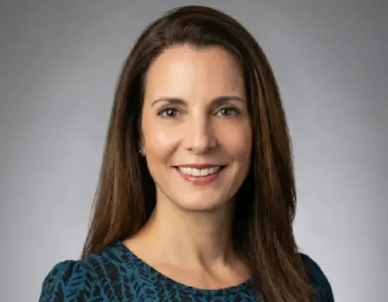 Headshot of a person with long brown hair smiling against a plain gray background, wearing a dark patterned top.