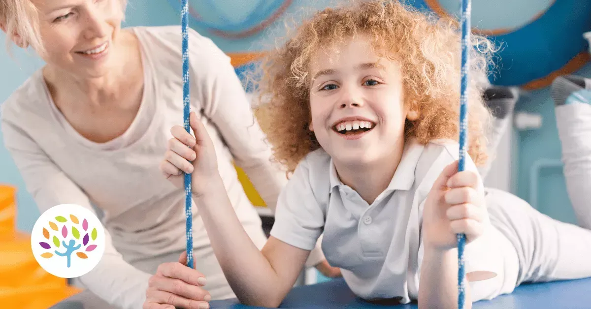 A person in a light shirt supervises a smiling child holding onto ropes while lying on a blue sensory swing.